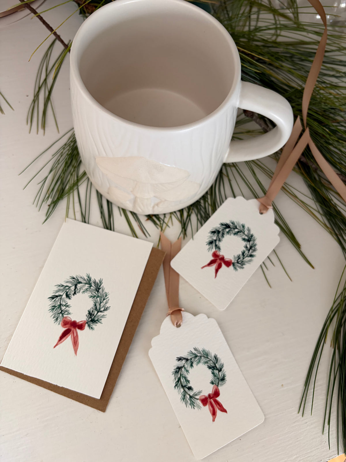 A white ceramic mug with a ribbed texture and a white leaf design on the side, styled with pine branches and two white gift tags featuring a green wreath and red bow, alongside a kraft envelope.
