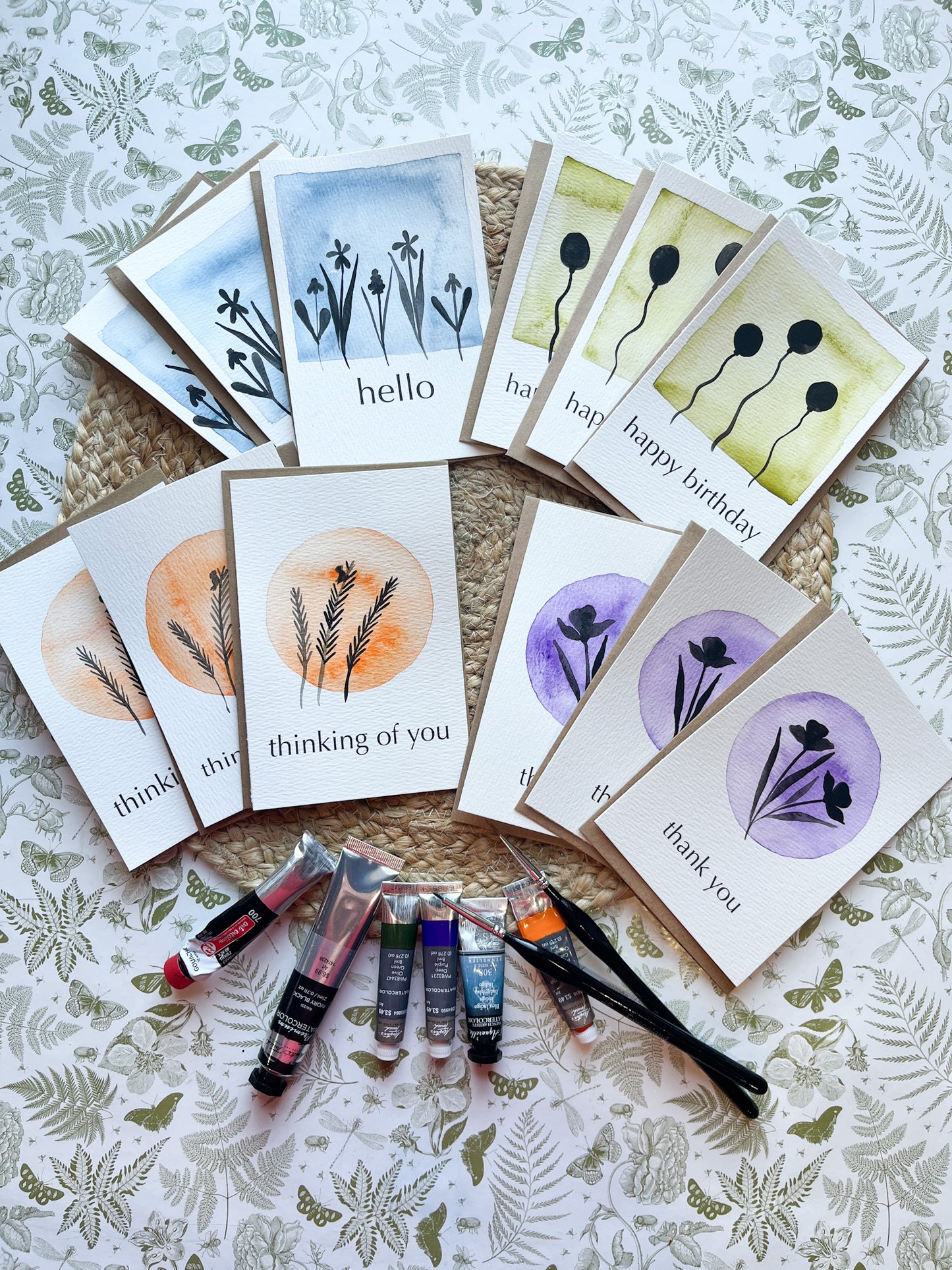Collection of greeting cards with floral designs and matching paint tubes on a floral-patterned surface.