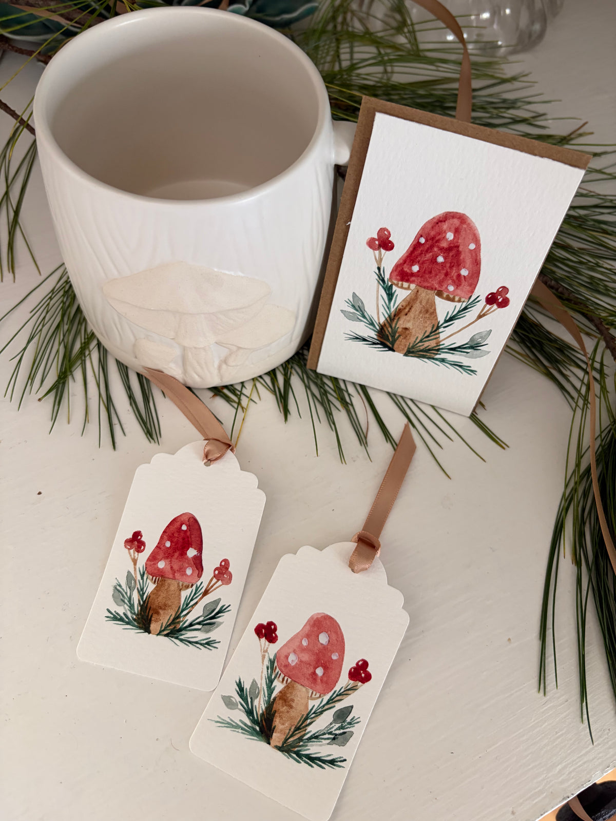 A white ceramic mug and two matching gift tags or cards decorated with a red-and-white mushroom illustration, arranged with pine branches on a light surface.