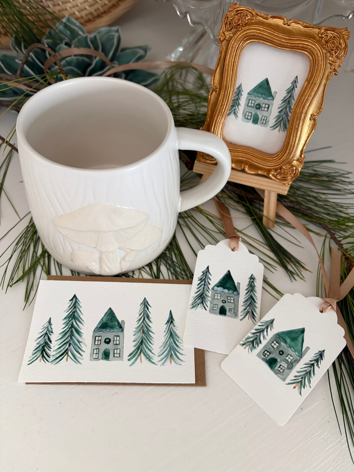 A white ceramic mug with a raised mushroom design sits beside a small gold-framed print, a matching card, and two gift tags, all decorated with a green watercolor house and pine tree motif on a styled tabletop with evergreen branches.