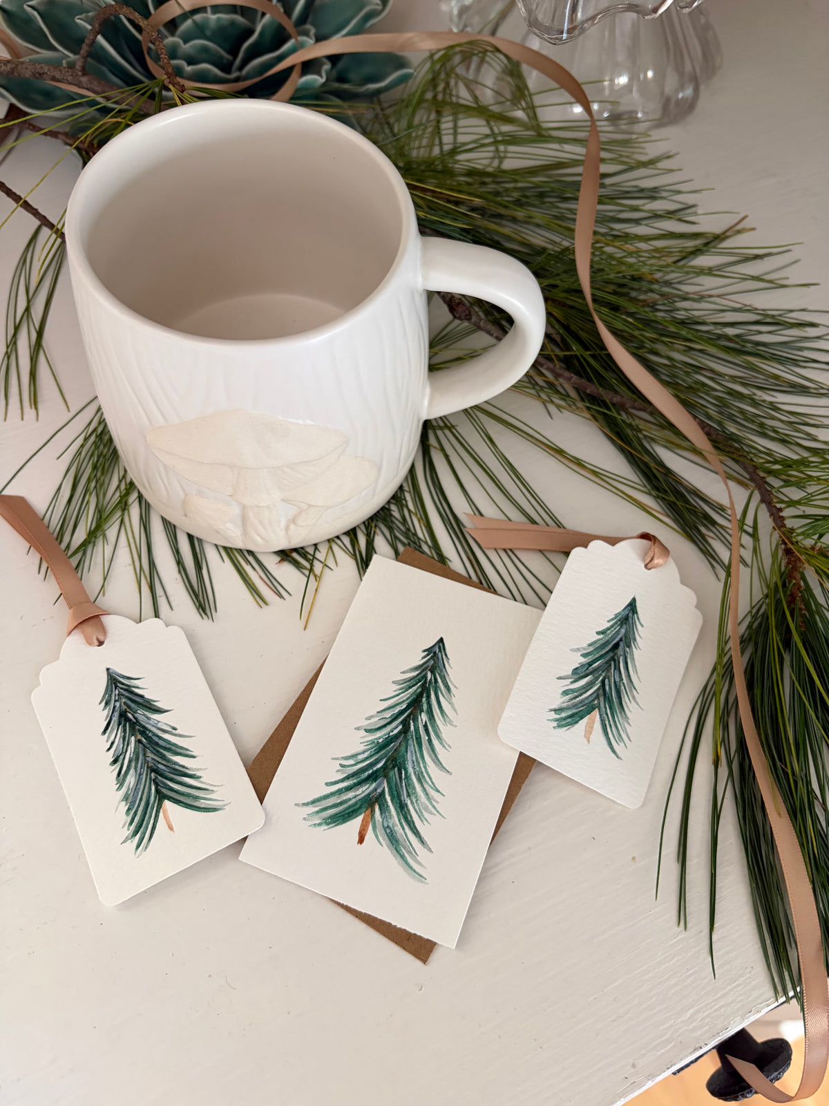 A white ceramic mug with a ribbed texture and a hand-painted green pine tree on the side, styled on a white tabletop with pine needles, brown ribbons, and matching gift tags.